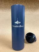 A matte navy blue insulated tumbler with a white Wooden Boat logo and text on the front, shown standing upright on a tan textured surface with its black lid placed beside it.