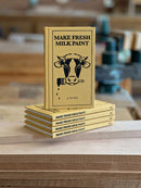 Stack of books titled 'Make Fresh Milk Paint' on a wooden surface with a blurred background.